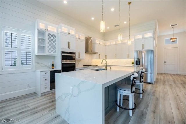 kitchen with stainless steel appliances, beverage cooler, wall chimney range hood, white cabinetry, and an island with sink