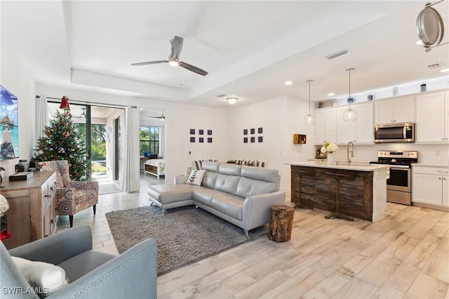 living room with ceiling fan, a tray ceiling, sink, and light wood-type flooring