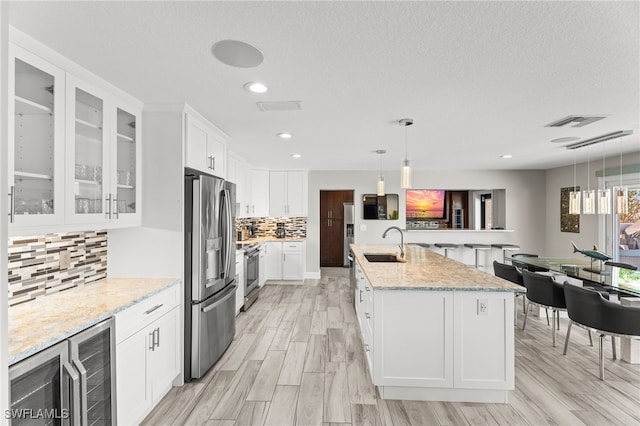 kitchen with white cabinets, wine cooler, an island with sink, appliances with stainless steel finishes, and decorative light fixtures