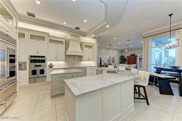 kitchen with stainless steel double oven, tasteful backsplash, a large island with sink, decorative light fixtures, and custom range hood