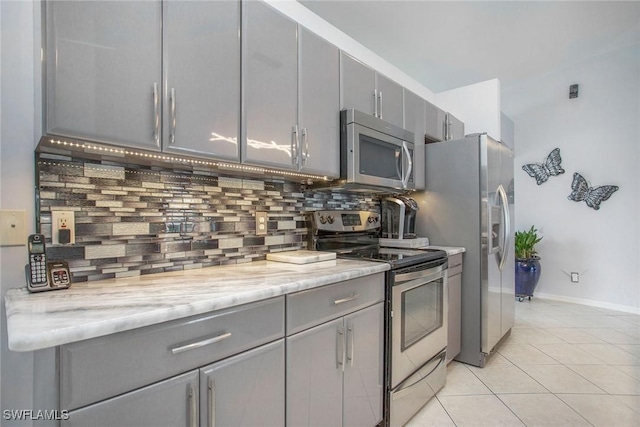 kitchen with gray cabinetry, backsplash, appliances with stainless steel finishes, light tile patterned flooring, and light stone counters