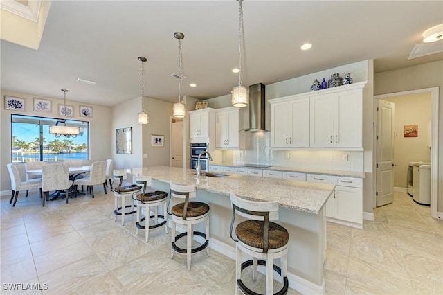 kitchen with hanging light fixtures, wall chimney exhaust hood, an island with sink, and white cabinets