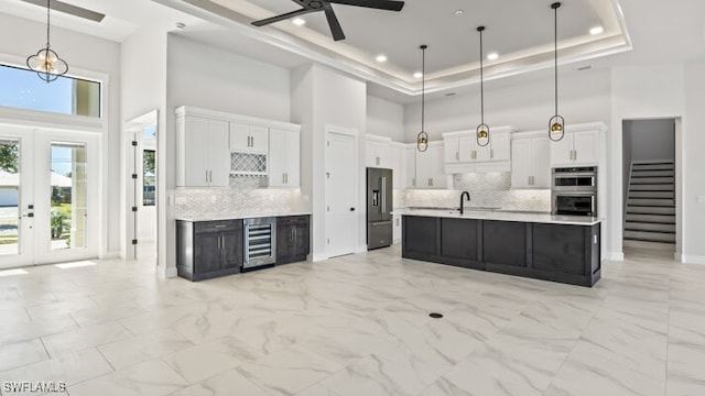 kitchen with a towering ceiling, a raised ceiling, pendant lighting, a center island with sink, and white cabinetry