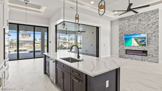 kitchen featuring ceiling fan, sink, a raised ceiling, a stone fireplace, and light stone counters