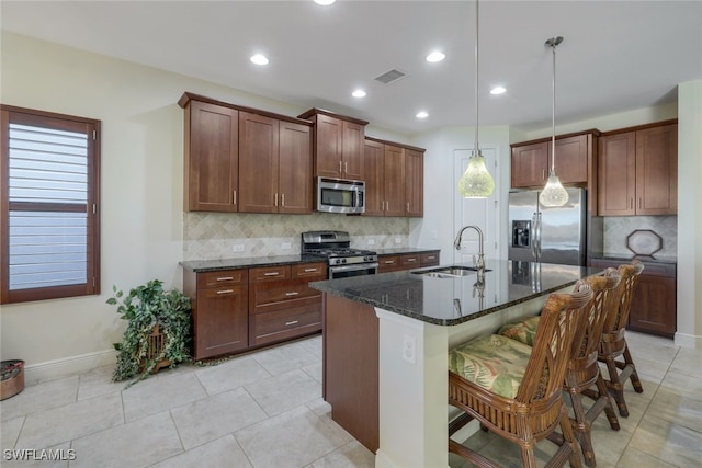 kitchen with pendant lighting, appliances with stainless steel finishes, a kitchen island with sink, a sink, and dark stone countertops