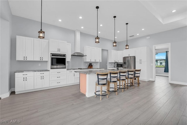 kitchen featuring wall chimney exhaust hood, stainless steel appliances, a kitchen island with sink, white cabinetry, and a kitchen breakfast bar