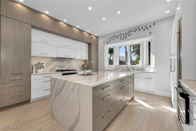 kitchen featuring a center island with sink, sink, decorative backsplash, light stone countertops, and white cabinetry