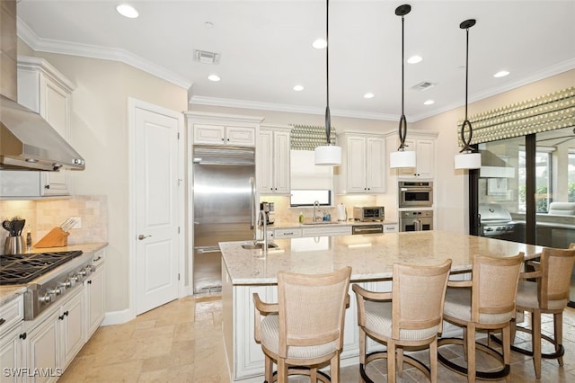 kitchen featuring light stone countertops, an island with sink, range hood, appliances with stainless steel finishes, and decorative light fixtures