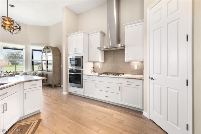 kitchen featuring wall chimney range hood, decorative light fixtures, light stone counters, white cabinetry, and stainless steel appliances