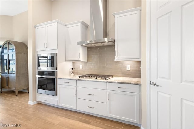 kitchen featuring wall chimney range hood, light stone countertops, light wood-type flooring, white cabinetry, and stainless steel appliances