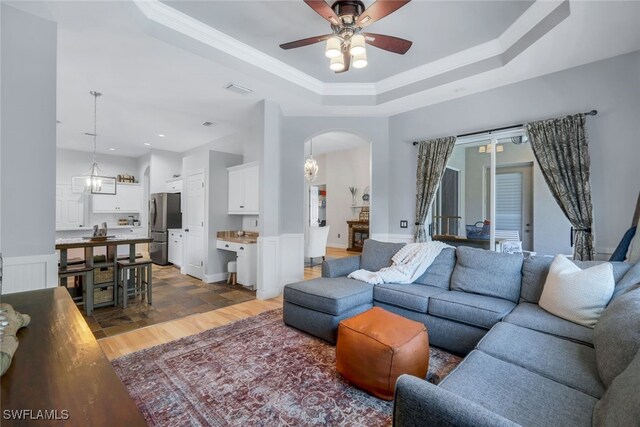 living room with arched walkways, crown molding, a raised ceiling, light wood-type flooring, and ceiling fan with notable chandelier