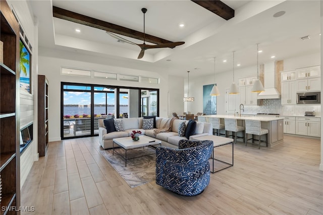 living room featuring sink, ceiling fan, light wood-type flooring, a tray ceiling, and beam ceiling