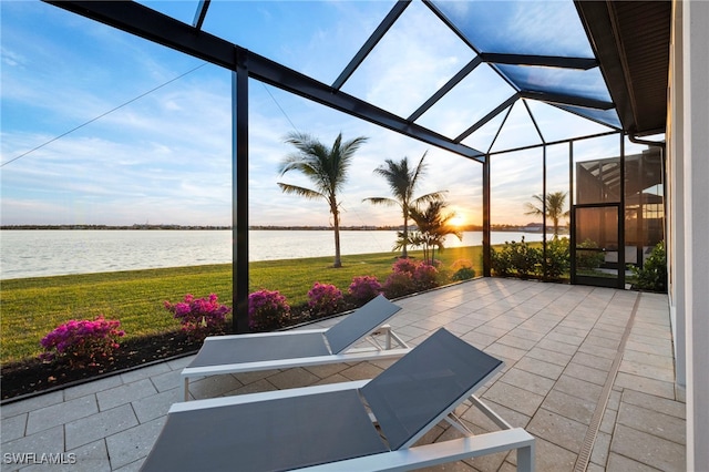 patio terrace at dusk featuring glass enclosure, a water view, and a yard