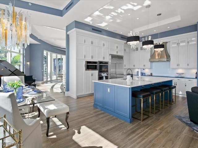 kitchen featuring appliances with stainless steel finishes, wall chimney range hood, a center island with sink, an inviting chandelier, and hanging light fixtures