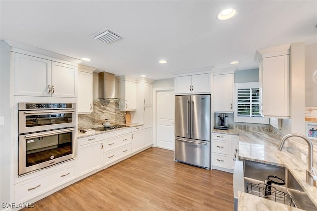 kitchen with light hardwood / wood-style flooring, wall chimney exhaust hood, light stone countertops, appliances with stainless steel finishes, and white cabinetry