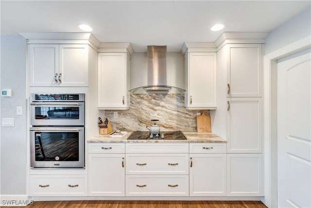 kitchen with wall chimney range hood, light stone counters, double oven, black electric stovetop, and white cabinets