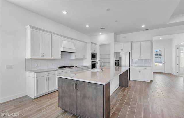 kitchen featuring custom exhaust hood, a kitchen island with sink, white cabinets, sink, and appliances with stainless steel finishes