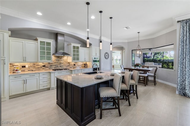 kitchen featuring sink, wall chimney range hood, light stone counters, pendant lighting, and a kitchen island with sink