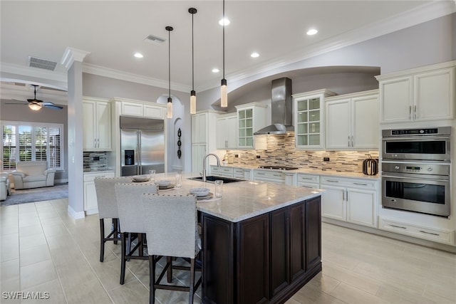 kitchen featuring sink, wall chimney exhaust hood, stainless steel appliances, an island with sink, and pendant lighting