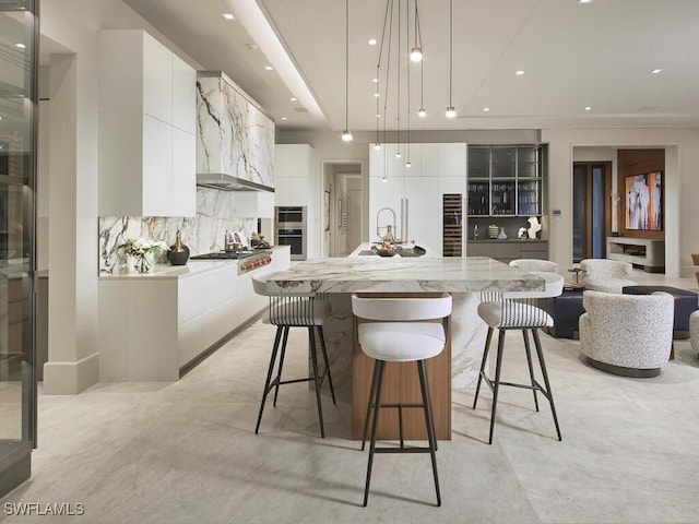 kitchen featuring wall chimney exhaust hood, decorative light fixtures, a kitchen island with sink, decorative backsplash, and white cabinets