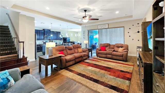 living area featuring a tray ceiling, ceiling fan, wood finished floors, baseboards, and stairs