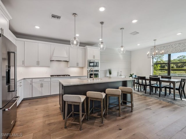 kitchen featuring hanging light fixtures, stainless steel appliances, a kitchen island with sink, white cabinets, and custom exhaust hood