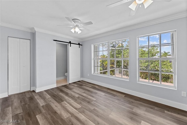 unfurnished bedroom with ceiling fan, a barn door, crown molding, wood-type flooring, and a closet