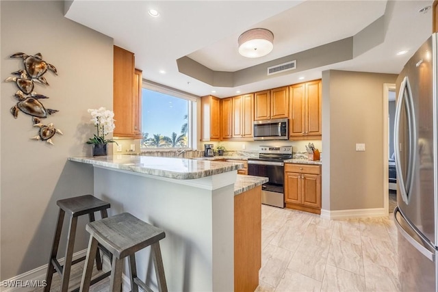 kitchen with kitchen peninsula, a kitchen breakfast bar, light stone countertops, stainless steel appliances, and a tray ceiling