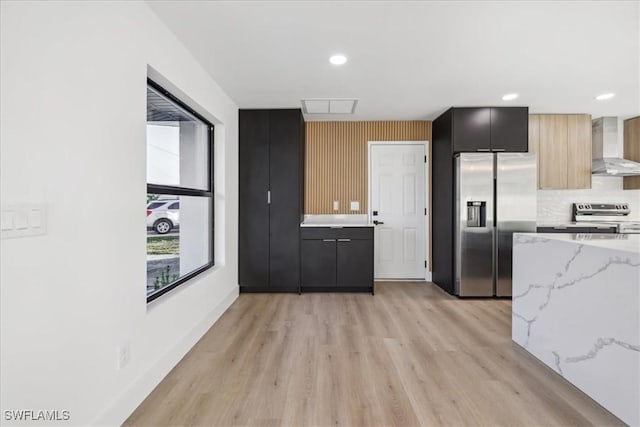 kitchen with wall chimney exhaust hood, light hardwood / wood-style flooring, appliances with stainless steel finishes, light stone countertops, and decorative backsplash