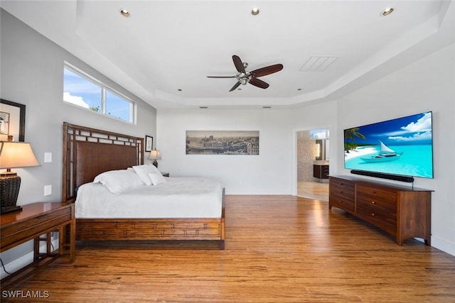 bedroom featuring ceiling fan, ensuite bathroom, light hardwood / wood-style flooring, and a tray ceiling