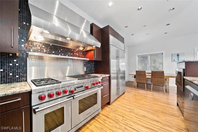 kitchen featuring built in appliances, wall chimney exhaust hood, light hardwood / wood-style floors, and tasteful backsplash