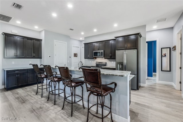 kitchen featuring appliances with stainless steel finishes, a breakfast bar, light stone countertops, a center island with sink, and light hardwood / wood-style flooring