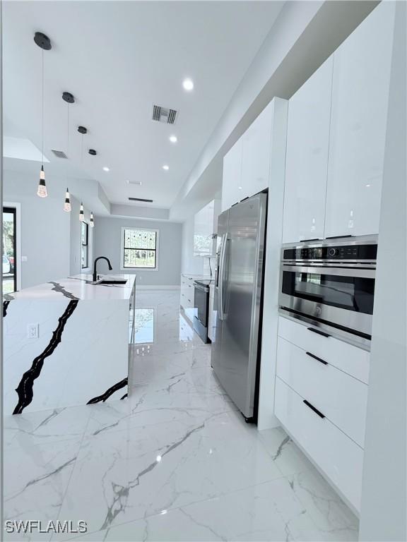 kitchen with appliances with stainless steel finishes, a kitchen island with sink, sink, white cabinets, and hanging light fixtures