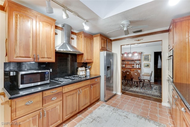 kitchen featuring wall chimney exhaust hood, dark stone countertops, appliances with stainless steel finishes, ceiling fan, and backsplash