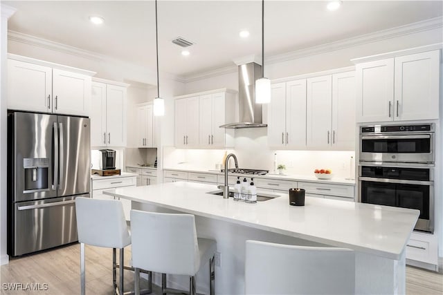 kitchen featuring hanging light fixtures, wall chimney range hood, an island with sink, white cabinets, and appliances with stainless steel finishes