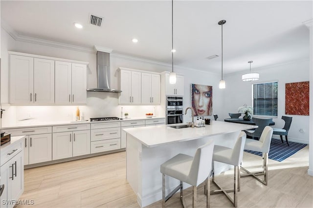 kitchen featuring a kitchen island with sink, wall chimney range hood, hanging light fixtures, a kitchen bar, and white cabinetry