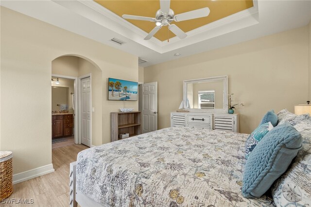 bedroom with a raised ceiling, ensuite bath, light hardwood / wood-style flooring, and ceiling fan