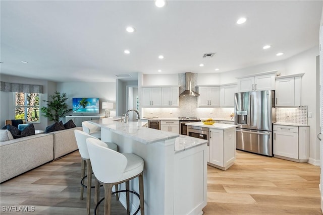 kitchen with an island with sink, white cabinetry, wall chimney exhaust hood, stainless steel appliances, and light hardwood / wood-style flooring