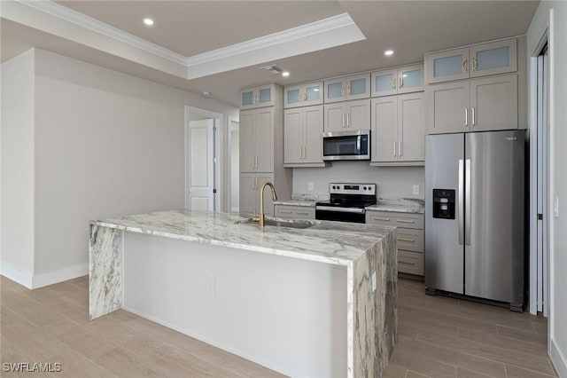 kitchen featuring light stone countertops, sink, a raised ceiling, an island with sink, and appliances with stainless steel finishes