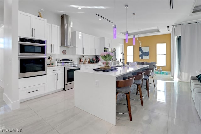 kitchen featuring a kitchen island with sink, white cabinets, wall chimney range hood, decorative light fixtures, and stainless steel appliances