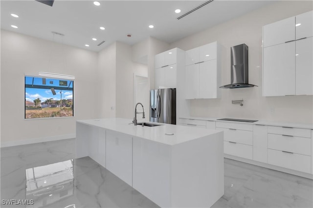 kitchen with an island with sink, stainless steel refrigerator with ice dispenser, white cabinets, wall chimney range hood, and sink