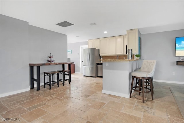 kitchen with kitchen peninsula, a kitchen breakfast bar, light stone countertops, cream cabinetry, and stainless steel refrigerator