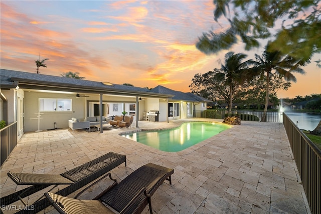 pool at dusk featuring outdoor lounge area, grilling area, a water view, ceiling fan, and a patio area