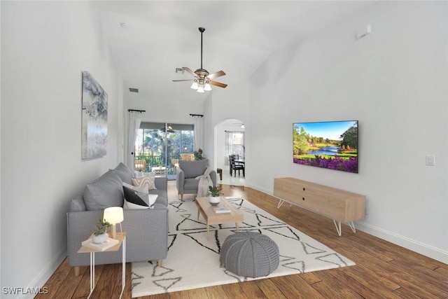 living room featuring wood finished floors, baseboards, high vaulted ceiling, arched walkways, and ceiling fan