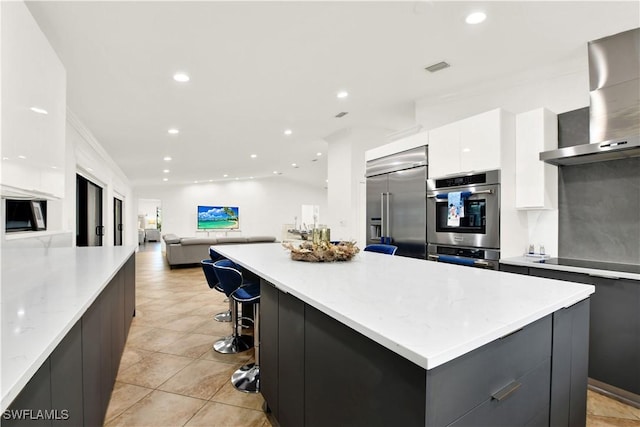 kitchen with appliances with stainless steel finishes, wall chimney exhaust hood, a breakfast bar, white cabinets, and a center island