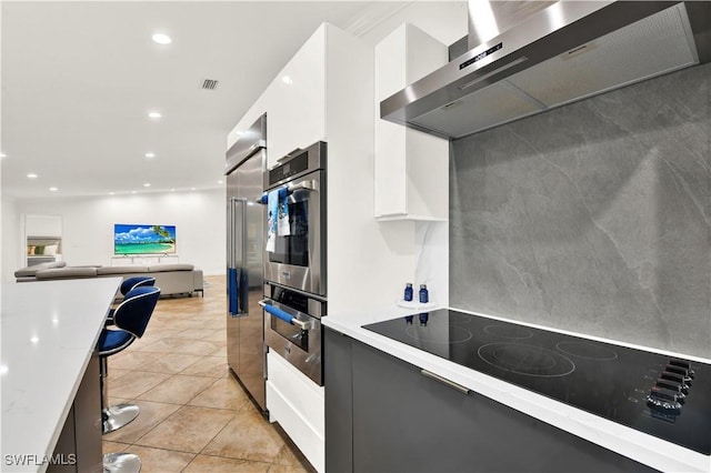 kitchen with white cabinets, black electric stovetop, wall chimney exhaust hood, light tile patterned floors, and stainless steel double oven