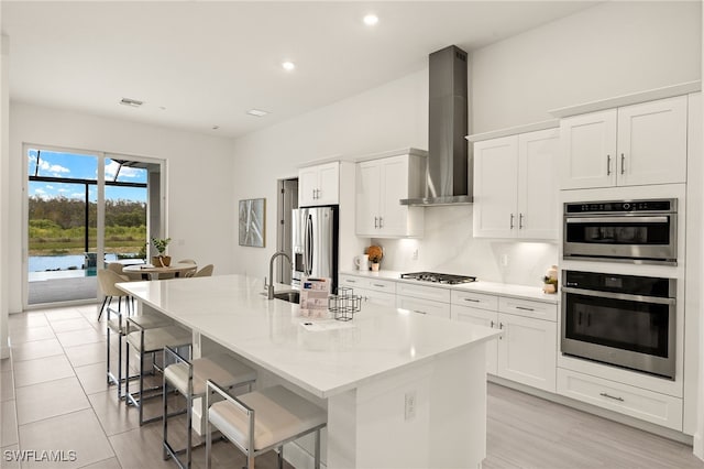 kitchen featuring stainless steel appliances, a kitchen bar, white cabinetry, an island with sink, and wall chimney exhaust hood