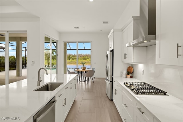 kitchen featuring appliances with stainless steel finishes, white cabinets, a sink, light stone countertops, and wall chimney exhaust hood