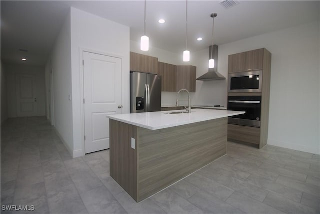 kitchen with appliances with stainless steel finishes, wall chimney exhaust hood, a kitchen island with sink, sink, and decorative light fixtures