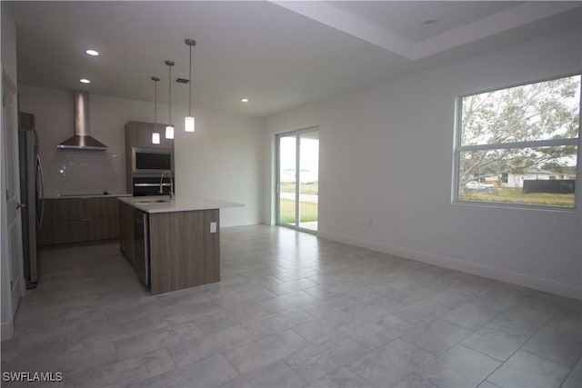 kitchen with a wealth of natural light, a center island with sink, wall chimney exhaust hood, and decorative light fixtures